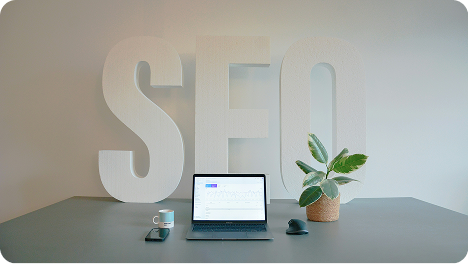 A well-lit desk setup in Johor Bahru, Malaysia, featuring SEO-related text on a background, a coffee mug, an iPad, a MacBook, and a small plant.